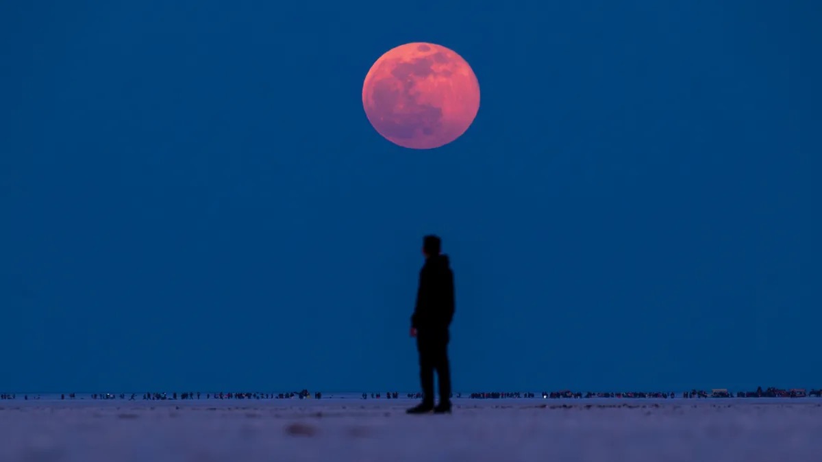 A rose-red moon above a distant silhouette beneath a wide blue sky.